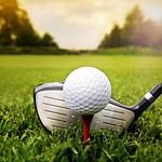 Golf ball on tee with club poised for swing on grass.