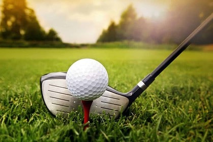 Golf ball on tee with club poised for swing on grass.