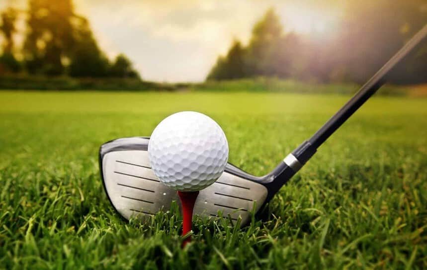 Golf ball on tee with club poised for swing on grass.