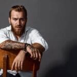 Handsome man with tattoos wearing white shirt sitting on a chair.