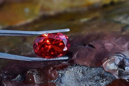Tweezers holding a red gemstone, possibly ruby, against a natural backdrop.