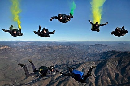 Skydivers in formation with colorful smoke trails over landscape.