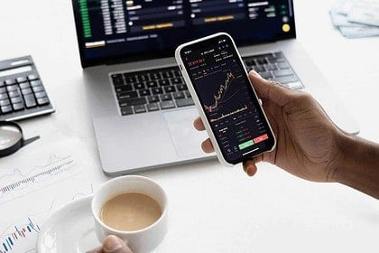 Person checking stock market on smartphone with laptop and coffee.
