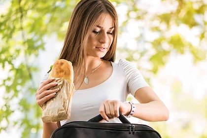 Busy woman eating sandwich, checking watch, holding briefcase outdoors.
